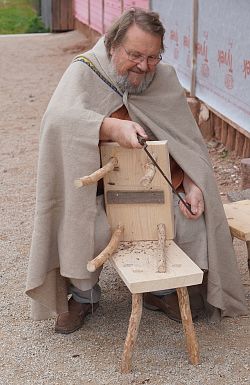 Dr. Luzian Taeger, gekleidet in ein römisches Gewand, bearbeitet einen Hocker nach römischen Vorbild mit einem Handhobel.