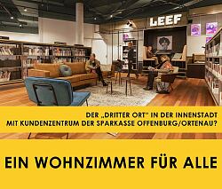 Zu sehen ist ein Raum mit Bücherregalen an den Wänden. Davor stehen ein Sofa und zwei Sessel sowie 3 kleine runde Tische auf einem Teppich. Im Hintergrund ist eine Leseecke mit Polstern in der Wand integriert. Im Raum befinden sich 4 Personen, 3 davon sitzen auf den Möbeln und lesen; eine Person läuft durch den Raum.