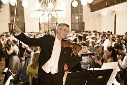 Ein Geiger steht in einem Ballsaal im Smoking, dahinter tanzende Paare. 