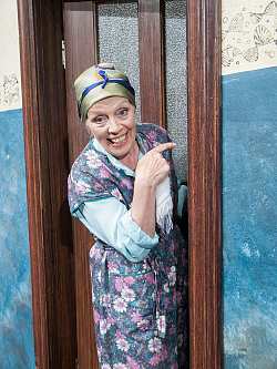 Zu sehen ist die Schauspielerin Heidi Mahler in ihrer Roller als Oma Dora. Sie trägt ein geblümtes Schürzenkleid in Lila- und Blautönen, sowie ein grünes Kopftuch. Sie steht in einer Tür, lacht und zeigt mit dem Finger zur Tür.