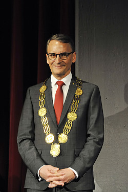 Zu sehen ist der neue Oberbürgermeister Markus Ibert bei seiner offiziellen Amtseinführung im Parktheater Lahr. Er steht auf der Bühne, hinter ihm der dunkelrote Bühnenvorhang. Er trägt einen grauen Anzug und eine rote Kravatte. Die goldene Amtskette trägt er um den Hals.
