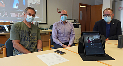 Die Drei sitzen an einem Tisch, im Hintergrund ist die App mit einem Beamer an die Leinwand projeziert , im Vordergrund steht ein IPad mit der Erna-App auf dem Bildschirm. 