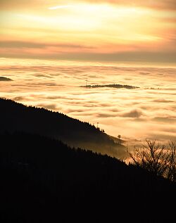 Blick von einem Berg über ein Wolkenfeld, Ein Windrad auf dem Rücken eine weiteren Berge schaut aus den Wolken, der Himmel ist in oranges Licht getaucht