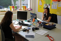 Das Bild zeigt zwei FSJ-lerinnen, die sich an zwei Schreibtischen mit PCs gegenüber sitzen. Die Linke schreibt, die rechte telefoniert.