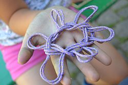 Auf der Innenfläche einer Kinderhand liegt eine Fadenspielfigur aus violetter Schnur.