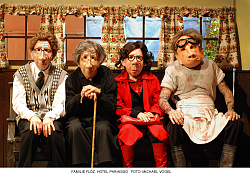 Szene aus dem Theaterstück "Familie Flöz - Hotel Paradiso" Vier Puppen sitzen auf einer Holzbank. Im Hintergrund ist eine Fensterfront zusehen. 