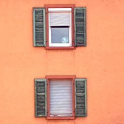 Hausfassade mit zwei Fenstern mit Fensterläden und Jalousien, eine davon ist geschlossen, die andere halb, darunter ist ein Ventilator zu sehen