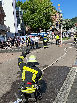 Feuerwehrmann der Stadt Lahr kniet auf einer Straße und dirigiert Einsatzkräfte, während mehrere Personen auf einem Gerüst eine Maßnahme durchführen. Zuschauer beobachten die Szene.