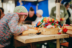 Kind mit bunter Mütze und Pullover sitzt an Holztisch und arbeitet mit Filzwolle und Werkzeugen