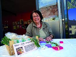 Frau Monika Fischer sitzt lächelnd hinter einem Tisch und hat vor sich mehrere verschiedenfarbige Seifen aufgebaut. 
