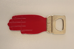 Es ist ein Flaschenöffner zu sehen, der die Form einer augestreckten Hand hat. Der Öffner bildet das Handgelenk. Die Hand selbst ist in dem spezifischen Rotton der Badischen Tabakmanufaktur gehalten.