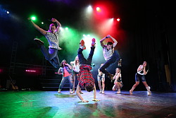 Tänzer zeigen vollen Einsatz und springen in die Luft, dahinter stehen Tänzerinnen die den Männern zuschauen.