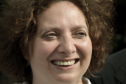 Portraitfoto von Claudia Toman. Sie blinzelt lachend in die Sonne und hat braunes lockiges Haar.