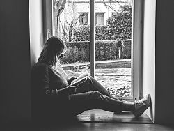 Das Foto ist schwarzweiß und zeigt eine junge Frau, die an einem Fenster sitzt und ein Buch liest.