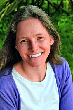 Portrait der Autorin Ronja Erb. Sie strahlt mit vielen Sommersprossen im Gesicht in die Kamera. Im Hintergrund grüne Pflanzen und Bäume. Sie trägt ein weißes T-Shirt und eine lila Strickjacke. 