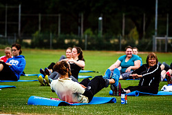 Auf einer grünen Wiese sitzen mehrere Personen unterschiedlichen Alters in Sportkleidung auf blauen Matten und machen Situps. 