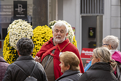 Ein Mann in roter Robe steht vor gelben Chrysanthemen und erzählt den Umstehenden etwas dazu.