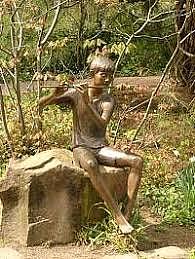 Metallskulptur, die eine Jungen zeigt, der Flöte spielt. Es sitzt auf einem Felsbrocken.