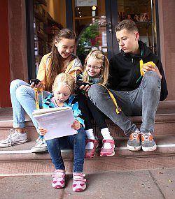Das Bild zeigt drei Mädchen links und einen Jungen rechts, die vor dem Alten Rathaus auf den Stufen sitzen und in ein Streckenbuch schauen. Die beiden älteren halten jeweils ein gelbes GPS-Gerät in der Hand.
