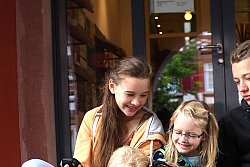 Das Bild zeigt drei Mädchen links und einen Jungen rechts, die vor dem Alten Rathaus auf den Stufen sitzen und in ein Streckenbuch schauen. Die beiden älteren halten jeweils ein gelbes GPS-Gerät in der Hand.