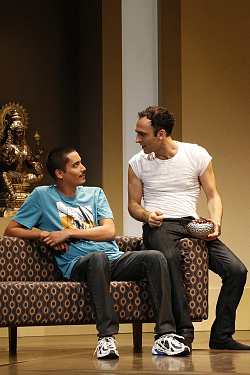 Zu sehen ist eine Szene aus dem Theaterstück "Geächtet". Zwei Männer sitzen auf einer Couch und seehn sich mit düsterer Miene an.