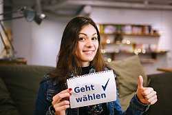 Das Bild zeigt eine junge Frau mit langen braunen Haaren. Sie trägt einen geblümten Schal und eine dunkelblaue Jeansjacke und lächelt in die Kamera. Ihren linken Daumen hält sie in die Luft gestreckt und in der rechten hand hält sie einen Zettel mit der Aufschrift "Geht wählen". Im Hintergund sieht man verschwommen eine Zimmereinrichtung mit einem Wandregal, einer Lampe und einer Couch.