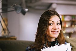 Das Bild zeigt eine junge Frau mit langen braunen Haaren. Sie trägt einen geblümten Schal und eine dunkelblaue Jeansjacke und lächelt in die Kamera. Ihren linken Daumen hält sie in die Luft gestreckt und in der rechten hand hält sie einen Zettel mit der Aufschrift &#34;Geht wählen&#34;. Im Hintergund sieht man verschwommen eine Zimmereinrichtung mit einem Wandregal, einer Lampe und einer Couch.