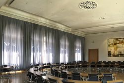 Auf dem Bild ist der Lahrer Gemeinderratssaal zu sehen. In der Mitte des Raumes sind kreisförmig in zwei Reihen die Tische und Stühle für die Gemeinderäte angeordnet. Der kreis ist nach vorne hin geöffnet. Dort stehen die Tische und Stühle für den Oberberbürgermeister und die Bürgermeister. Dahindet ist im Halbrund eine große Leinwand mit Beamern installiert. An der hinteren Wand gegenüber des Haupteingangs hängt ein großes vierteiliges modernes Kunstwerk an der Wand, rechts und links davon sind Türen. Davor sind die Tische und Stühle für die Verwaltungsmitarbeiter angeordnet. Links im Bild vor den großen Fenstern, die mit Vorhängen verschlossen sind, stehen die Tische für die Medienvertreter. Links von Haupteingang stehen die Besucherstühle. Die Tische sund jeweils mit Mikrofonen bestückt.