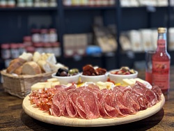 Runde Holzplatte mit aufgerollten Salami- und Schinkenaufschnitten, im Hintergrund Brot, kleine Schalen mit Oliven und weiteren Beilagen sowie eine Flasche mit rotem Getränk auf einem Holztisch.