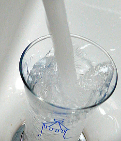 Ein Glas mit einem blauen Storchenturm steht in einem Waschbecken und wird von einem Strahl Wasser aus dem Wasserhahn gefüllt. 