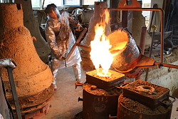 Ein Glockengießer fertigt die neue Bronzeglocke für Langenwinkel an. In einem Kessel über dem Feuer befindet sich flüssige Bronze, die in eine Form gegossen wird.