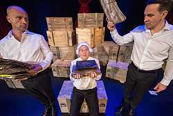 Theaterszene mit drei Männern, alle gekleidet mit schwarzen Hosen und weißen Hemden. Mann links hält mehrere vergilbte Blättersammlungen in den Händen und schaut bedrückt. Mann in der Mitte trägt weiße Perücke im Stil eines Richters, sitzt auf einer Holzkiste und hält ebenfalls Schriftsammlungen in den Händen. Mann rechts hält eine vergilbte Mappe mit Schriftstücken in die Höhe und spricht zu den beiden anderen. Im Hintergrund sind viele Holzkisten aufeinander gestapelt.