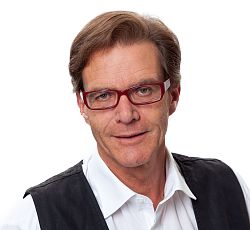 Portraitfoto von Gottfried Berger. Er hat braunes Haar und trägt eine rote Brille, außerdem ein weißes hemd mit einer schwarzen Weste.