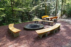 Halbrunde Sitzbänke aus Holz um einen runden Steinkreis als Feuerstelle in einem Waldgebiet