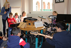 Um einen achteckigen Tisch mit einem Mischpult und Koopfhörer sitzen mehrere Kinder im Kreis. Drei probieren sich am Keyboard, ein Mädchen spielt Schlagzeug und zwei Jungs spielen E-Bass. Sie sieht man von hinten. Eine Lehrerin steht bei den Schülern am Keyboard und dirigiert. 