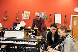 Im GrooveLab der städtischen Musikschule sitzt Marion Gentges neben einem Schüler mit E-Gitarre. Daneben bückt sich Tobias Meinen zu den Zweien hin. 