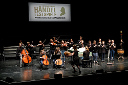 Das Barockorchester auf der Bühne. Andrea Cavalli steht mit einer Trommel davor. Die Musiker stehen uns spielen Streichinstrumente und Blockflöten. 