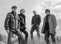 Vier Männer, jeweils mit Jeans und Jacken gekleidet, stehen auf einer Wiese mit hohem Gras. Zwei von ihnen tragen Mützen, einer zusätzlich eine Sonnenbrille.  Sie schauen mit ernster Miene in die Kamera. Das Bild ist schwarz-weiß.