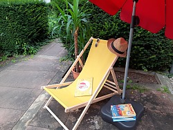 Auf dem Bild ist ein Liegestuhl in gelb mit Sonnenbrillen und Büchern zu sehen. Im Hintergrund befindet sich eine Pflanze.