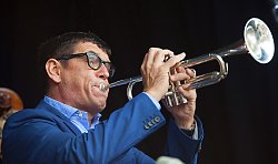 Zu sehen ist Helmut Dold, der in eine Trompete bläst. Er trägt ein blaues Jacket, ein hellblau-weiß kariertes Hemd und eine schwarze Brille