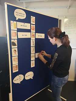 Eine Frau mit Pferdeschwanz steht vor einer Stellwand und heftet ein Stück Papier an. 