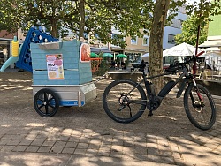 Fahrrad mit Anhänger, der eine blaue Holzkiste mit Aufklebern und einem roten Rücklicht trägt, im Park vor einem Spielplatz und Cafés:
