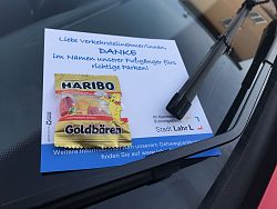 Das Bild zeigt die Frontscheibe eines roten Autos. Unter dem Scheibenwischer hängt ein weißes Kärtchen auf dem steht: &#34;Liebe Verkehrsteinhmer/innen, Danke im Namen unserer Fußgänger fürs richtige Parken. An dem Kärtchen wurde eine kleine Tüte Gummibärchen getackert. 