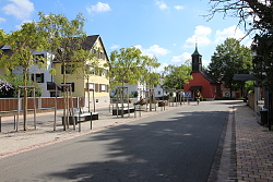 Das Bild zeigt die Wylerter Hauptstraße nach dem Umbau. In der Mitte ist ein Grünstreifen mit Bäumen, Bänken und einem Brunnen zu erkennen, auf der rechten Seite ein Bus-Wartehäuschen und im Hintergrund die rotgestrichene Kapelle.