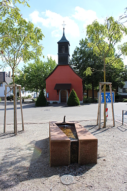 Das Bild zeigt die rotgestrichene St.-Blasius-Kapelle in Kippenheimweiler durch zwei Bäume links und rechts hindurch und über einen Brunnen hinweg.