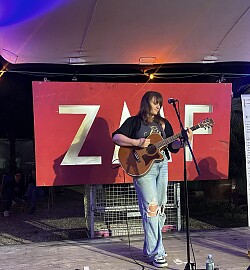Frau mit langem Haar in zerrissener Jeans spielt akustische Gitarre auf einer Bühne vor rotem Banner mit weißer Aufschrift.