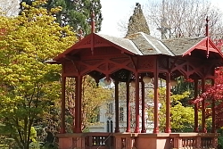 Sicht auf das rote Pavillon, daneben die schönen Bäume mit bunten Blättern, im Vorgrund eine weisse Besucherbank