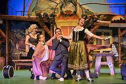 Frau im Trachtenkleid singt im Stehen und wird von drei Männern, einer im Anzug, die anderem ebenfalls in Tracht, umkreist und angehimmelt.