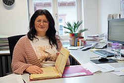 Das Bild zeigt Inna Vollenweider in ihrem Büro im Stadtplanungsamt Lahr. Inna Vollenweider hat dunkles langes gewelltes Haar und trägt eine Brille. Sie lächelt freundlich und schlägt gerade einen Bebauungsplan auf, der vor ihr auf dem Tisch liegt. Gekleidet ist sie mit einer rosafarbenen Strickjacke und einem weißen T-Shirt mit einem Aufdruck vorne.
