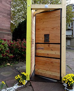 Installation vor der Kirche St. St. Peter und Paul und Lahr. Gezeigt wird eine große Holztür mit Chrysanthemenschmuck zu beiden Seiten. Über der Tür ist ein Lautsprecher angebracht. Ein Bewegungsmelder registriert, wenn jemand durch die Tür geht, so dass eine Stimme zu hören ist, die demjenigen Gedanken zum Glauben mit auf den Weg gibt.